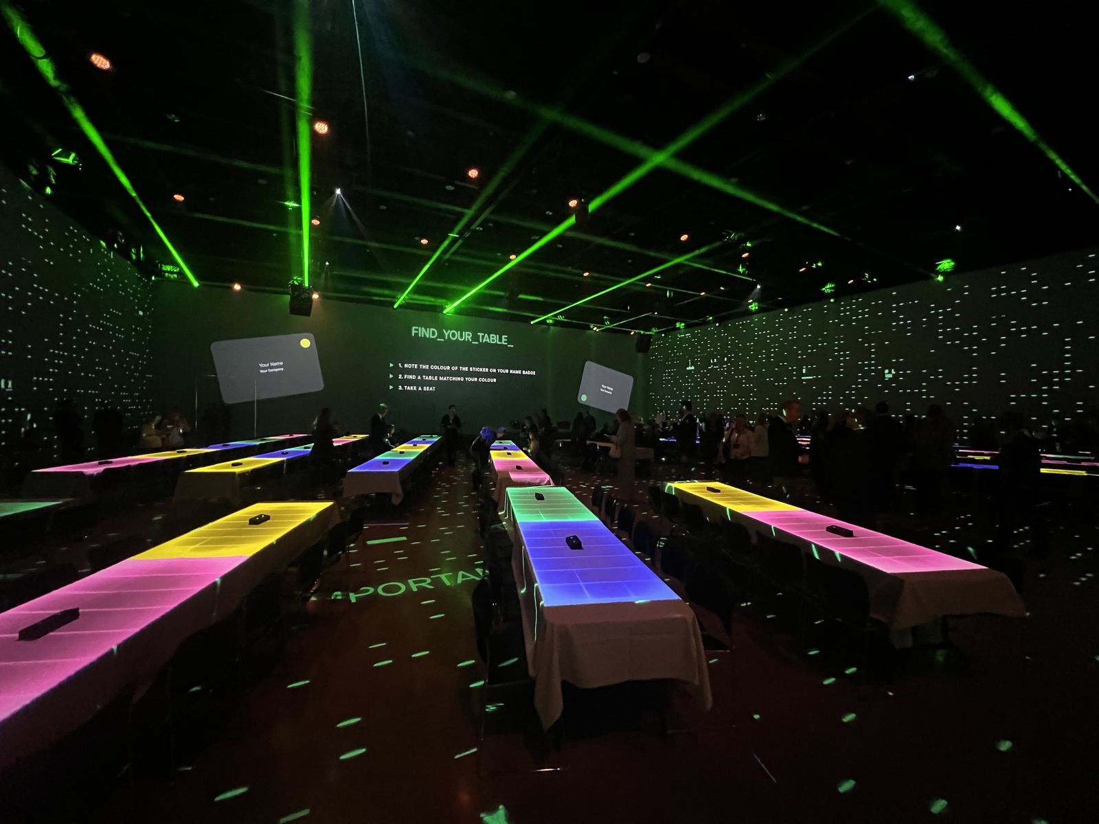En mörk eventsal med långbord i vita dukar. Varje bord lyser i sin egen färg från projektorerna i taket, gröna laserstrålar korsar rummet och på väggen står Find your table.